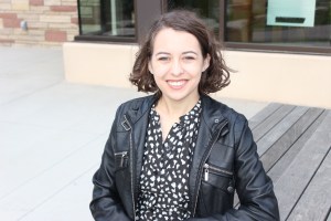 Woman sitting on bench in a black and white dress and a black jacket smiles at the camera
