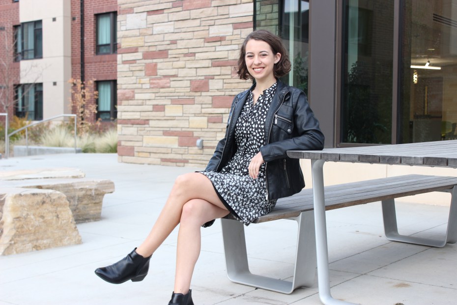 Woman sits on a picnic table bench in a black dress with black boots and a black jacket